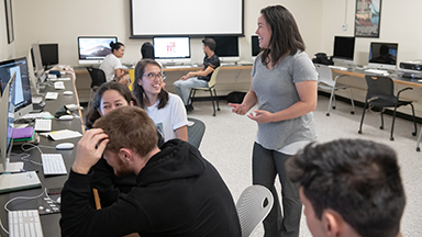 A professor smiles with students in a computer lab during a MAC class